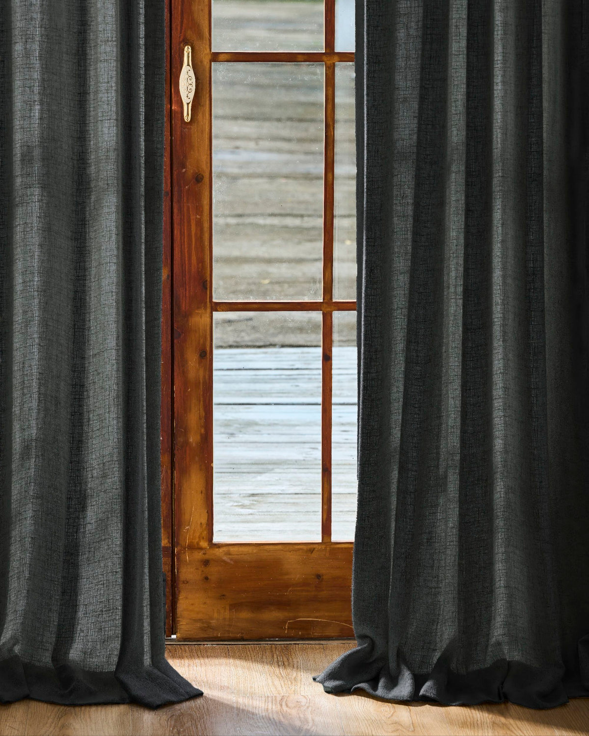 Dark grey linen blend custom curtains, richly textured, framing a wooden door with glass panes & puddling on the floor. High-end window treatments.