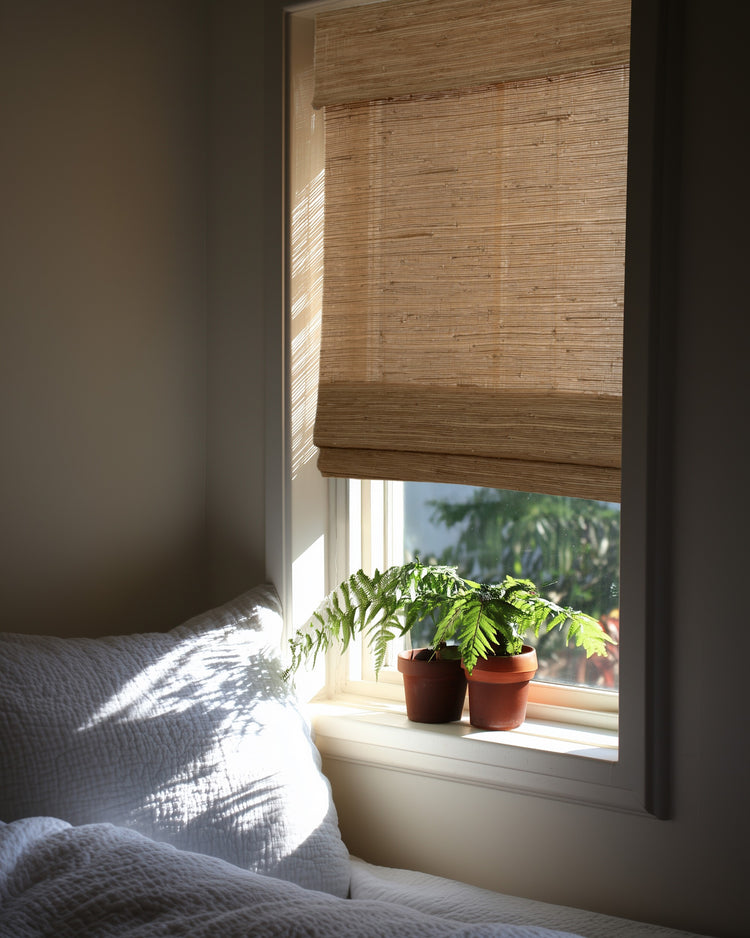Mia Jute Woven Shade in Sand covering a window, filtering sunlight onto a white bed and ferns. Provides natural light control & privacy.