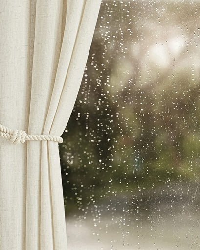 Light beige Noah linen-look waterproof custom outdoor curtain tied back with a rope, seen against a rain-splashed window.