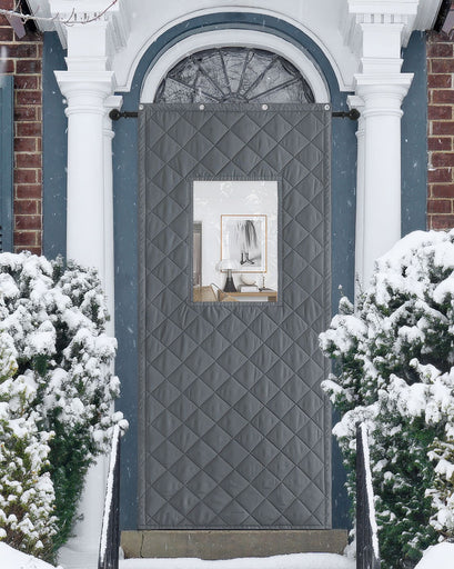 Gray quilted custom Oxford thermal door curtain with a PVC window on a snowy front door, providing insulation and soundproofing.