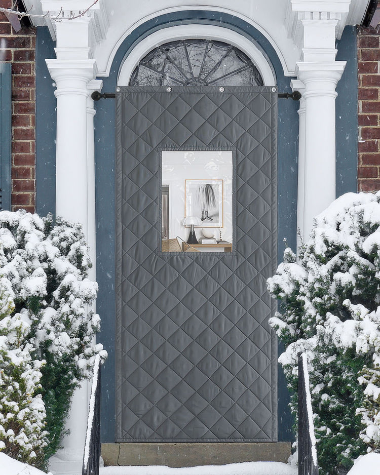 Gray quilted custom Oxford thermal door curtain with a PVC window on a snowy front door, providing insulation and soundproofing.