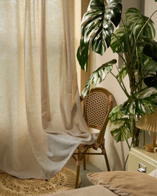 Light beige linen blend custom curtain with gentle transparency, draped beside a wicker chair and a large Monstera plant, enhancing natural home decor.