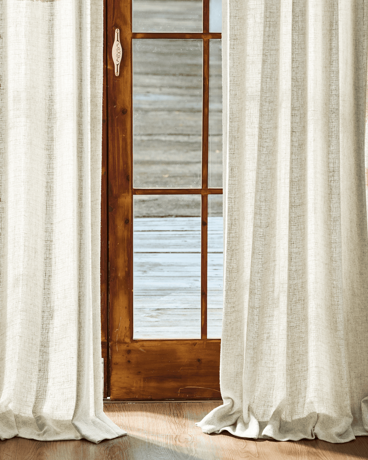 Textured Fiona Linen Blend custom curtains in light beige, framing a wooden glass door, pooling on a wooden floor. Elegant window treatment.