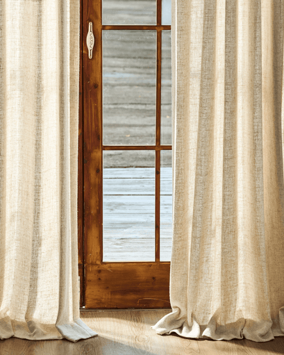 Fiona linen blend custom curtains in a natural shade frame a wooden door, puddled on the floor, showcasing light-filtering drapery.