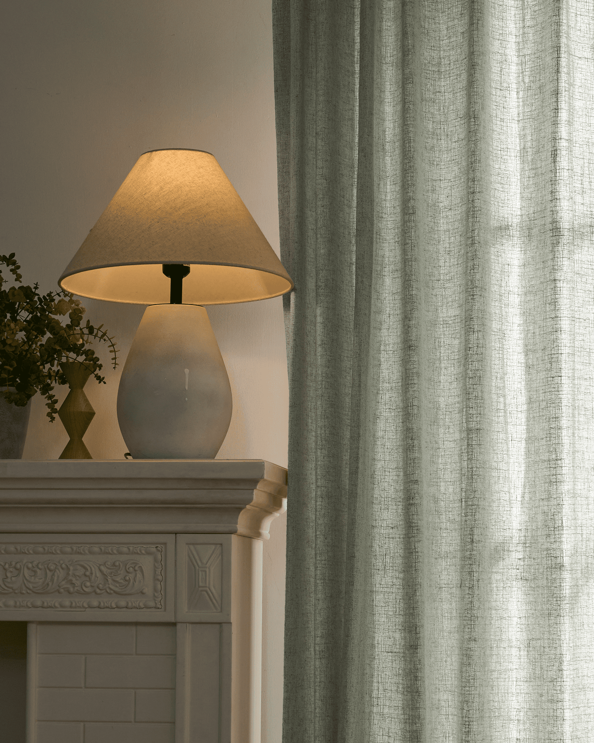 Textured light green Fiona linen blend custom curtain softly diffuses light; a glowing lamp sits on a white decorative mantel.