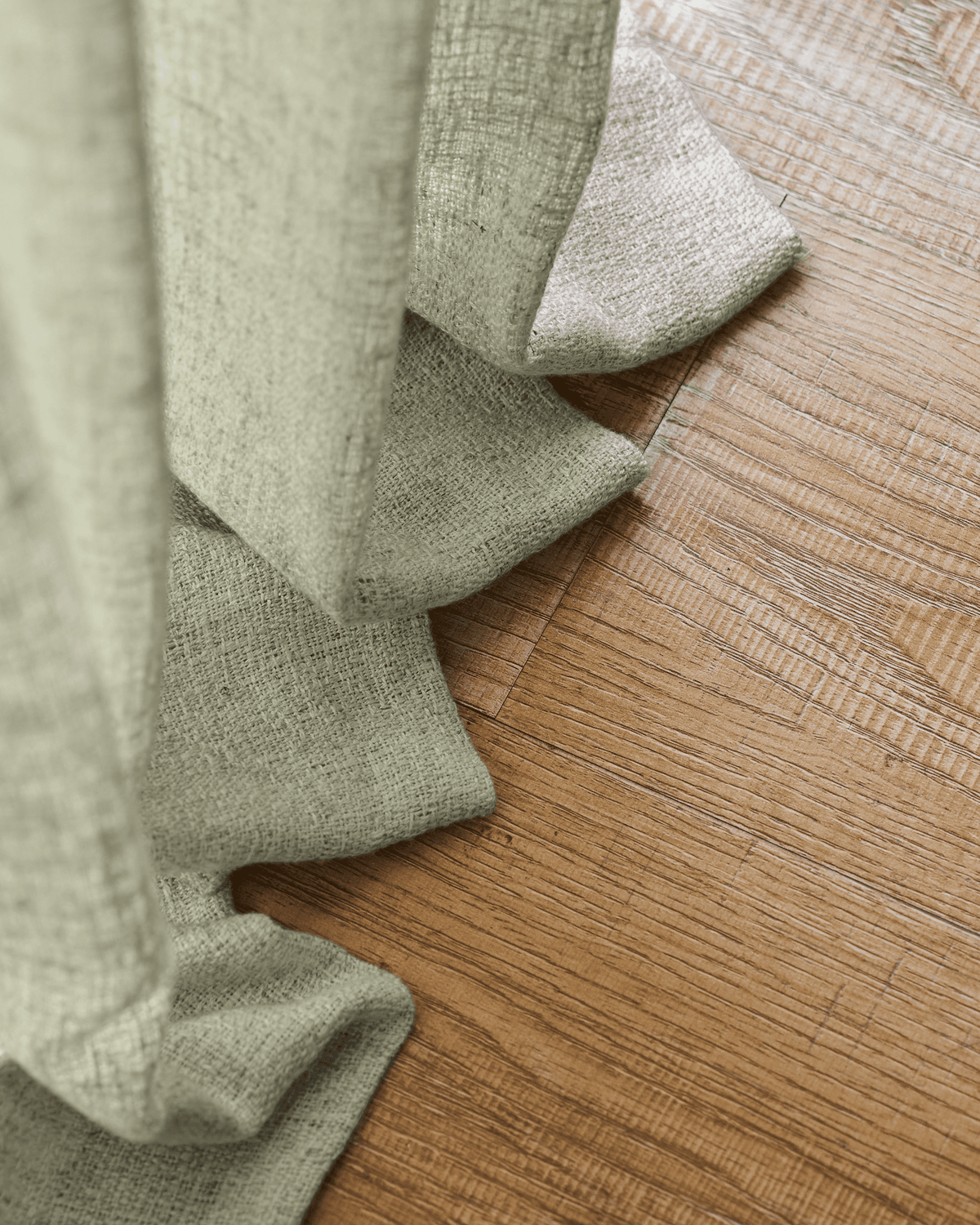 Textured Fiona Linen Blend custom curtain in soft green, elegantly folded on a warm hardwood floor, highlighting the luxurious fabric.