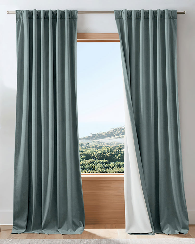 Custom dark teal velvet blackout curtains with pleated header, one panel revealing its white thermal lining for superior light control.
