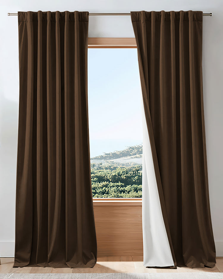 Custom brown velvet blackout curtains, one panel drawn back revealing its white thermal lining, perfectly framing a bright window view.
