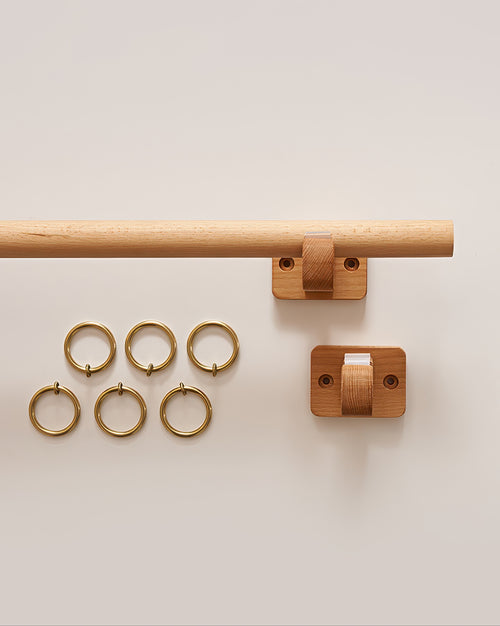 Minimal beech wood curtain rod with two matching wall mounts and six brass curtain rings for window treatments.
