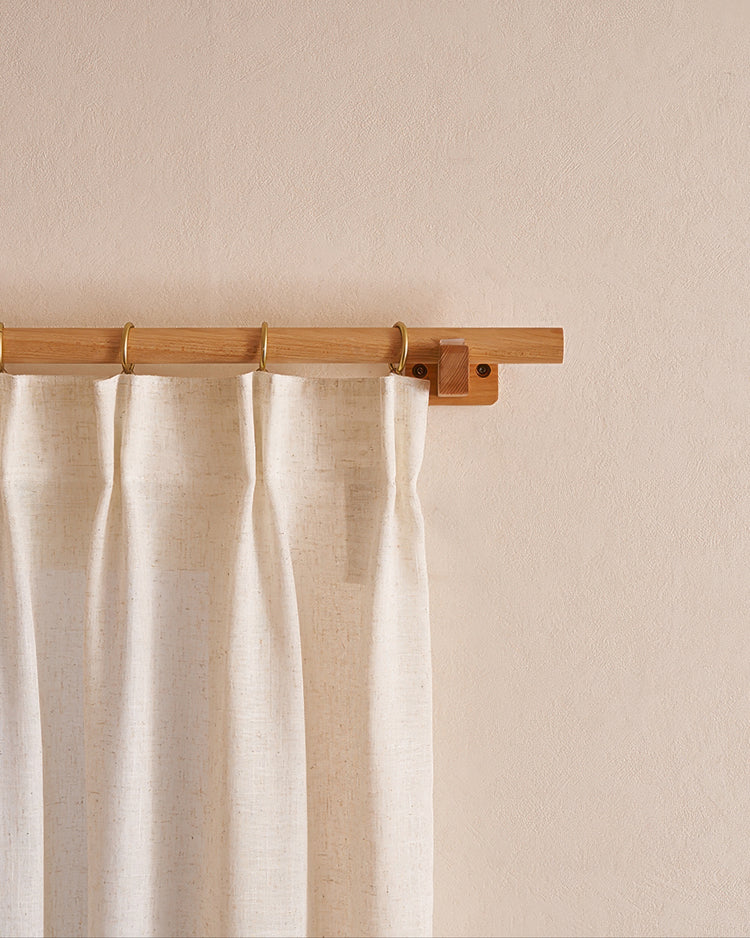 Close-up of a minimal beech wood curtain rod, brass rings, and a textured natural linen drape on a light wall. Custom window treatment hardware.