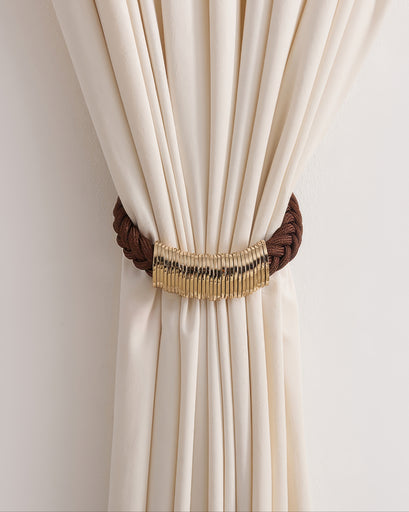 Brown braided ringed rope curtain tieback with gold metal detail cinching a soft cream drapery panel.