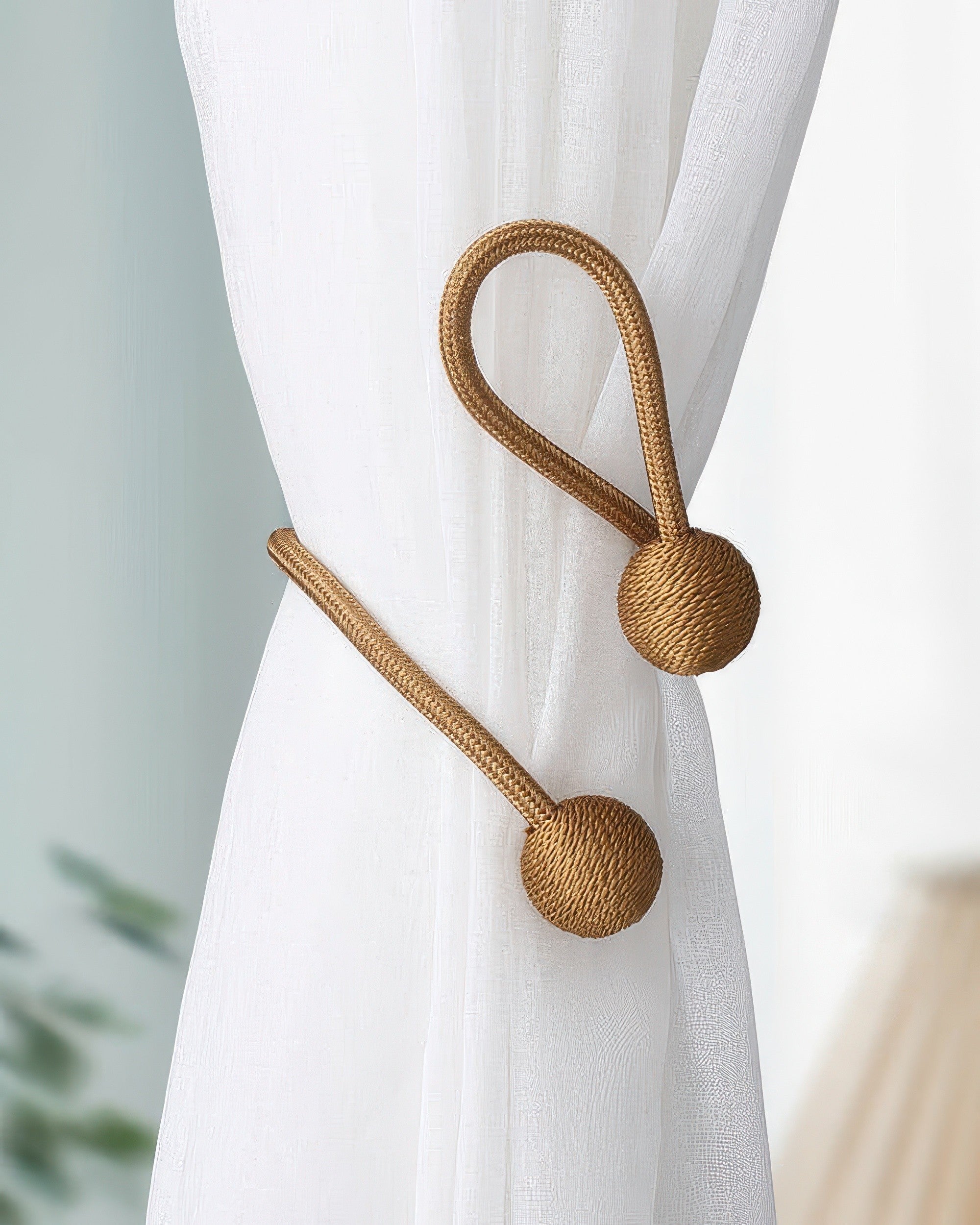 Elegant golden braided curtain tiebacks with spherical ends cinching sheer white drapery, perfect for sophisticated window treatments.
