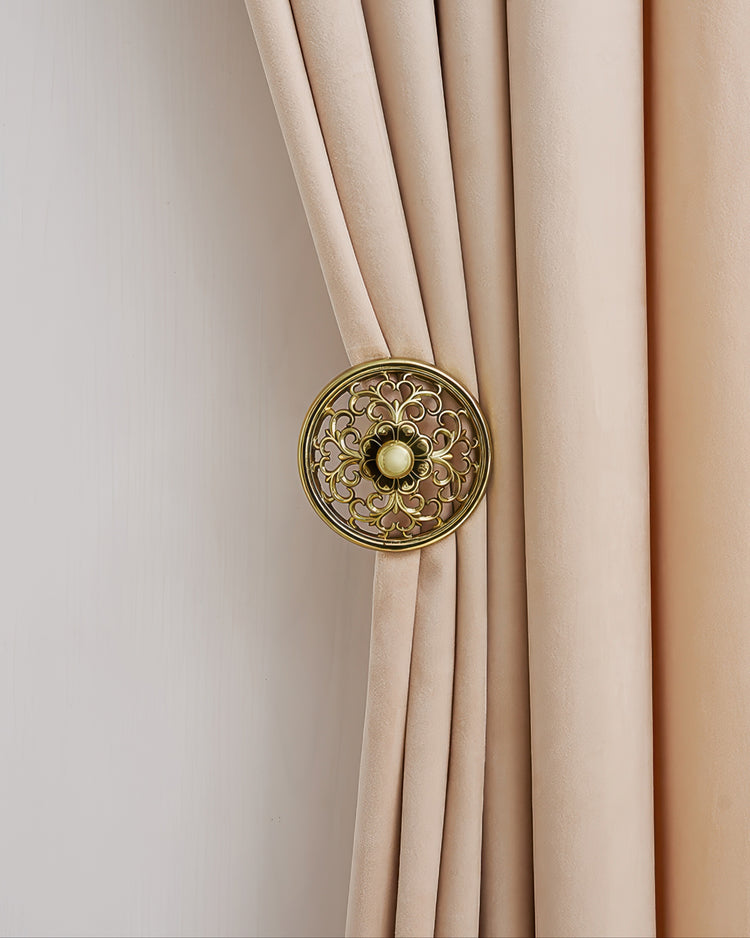 Ornate gold engraved curtain tie-back with central accent, securing luxurious pleated beige velvet drapery panel for custom window treatments.