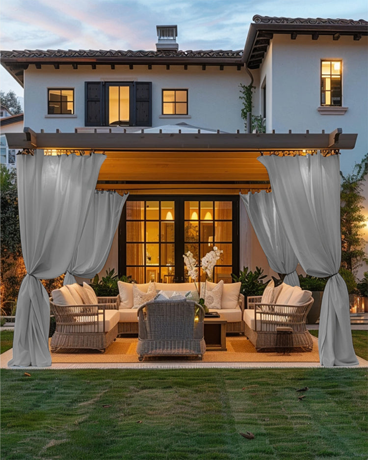 Custom light gray FadeShield Pro waterproof outdoor curtains tied back on a patio pergola with elegant seating at dusk.