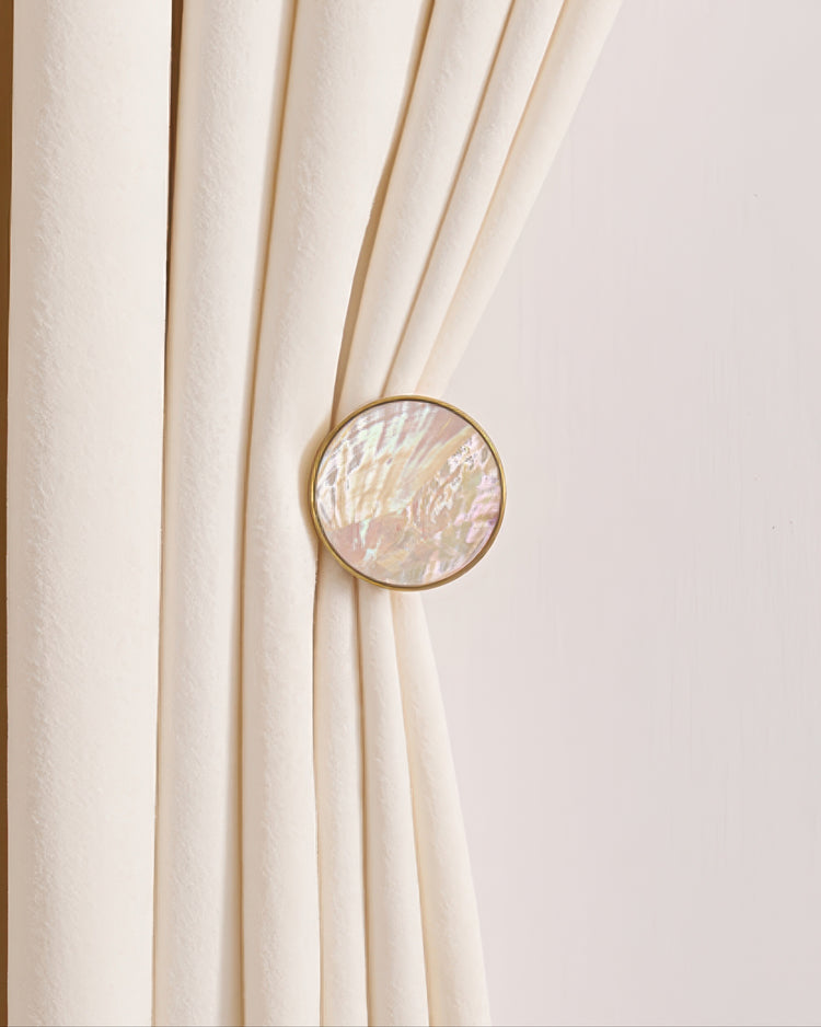 Elegant cream velvet drapery held by a round gold-framed seashell curtain wall hook for sophisticated window treatments.