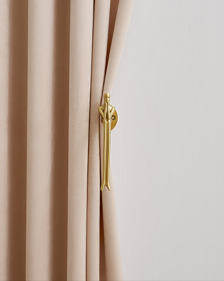 Close-up of a gold mannequin metal curtain hook holding back a flowing beige velvet drapery panel, accenting custom window treatments.