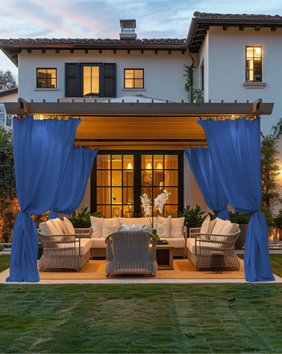 Blue FadeShield Pro™ custom outdoor curtains tied back on a pergola, providing privacy for a luxurious patio with wicker furniture at dusk.