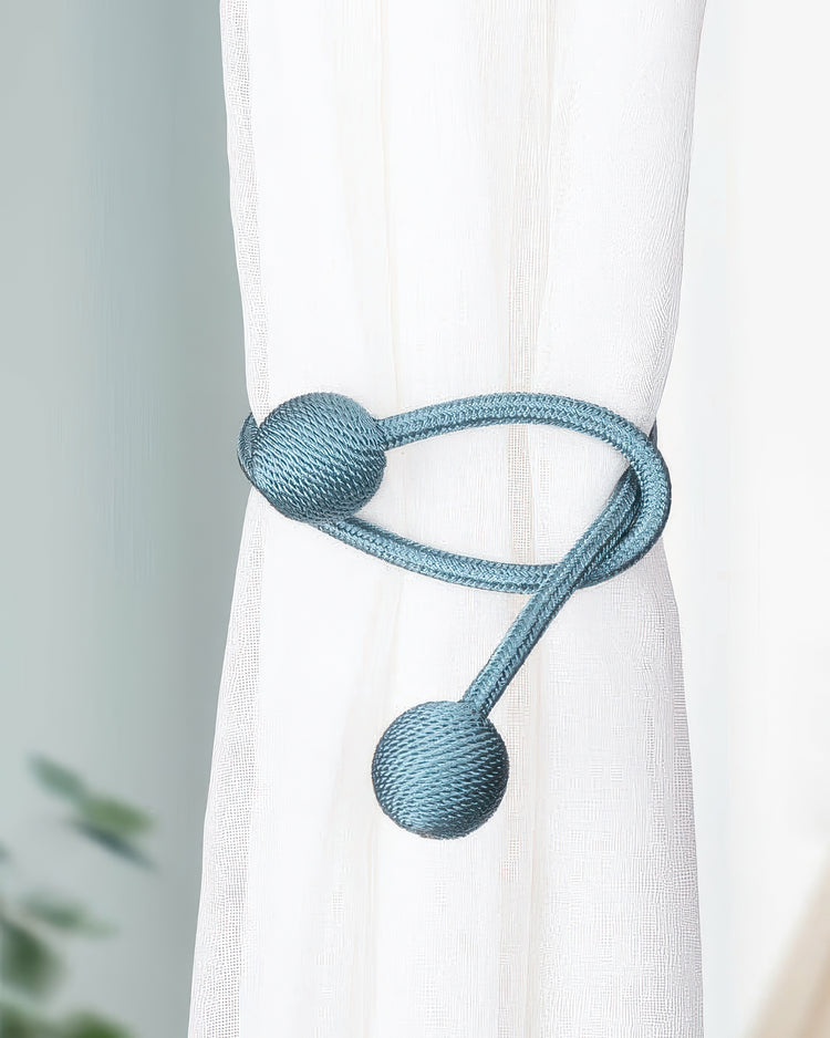 Teal braided rope curtain tieback with decorative spheres securing a white sheer drapery, enhancing modern window treatments.