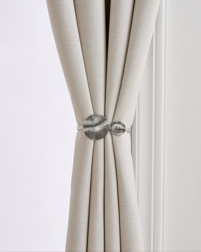 Elegant silver springed round snap curtain tieback with textured organic discs securing a luxurious light beige linen drapery.