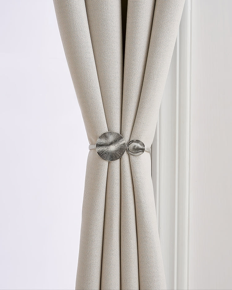 Elegant silver springed round snap curtain tieback with textured organic discs securing a luxurious light beige linen drapery.