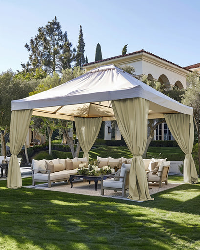 Elegant beige FadeShield™ canvas waterproof custom outdoor curtains tied back on a large patio gazebo with lounge seating & lush landscaping.