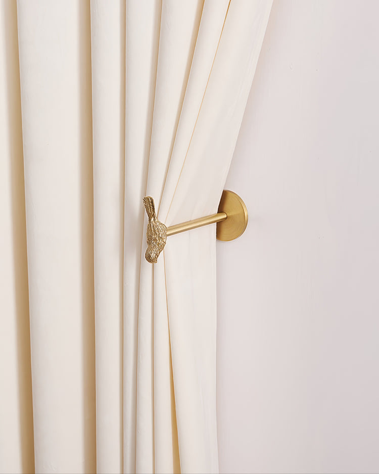 Textured gold bird metal curtain hook holding back a cream pleated luxury velvet drapery panel on a white wall.