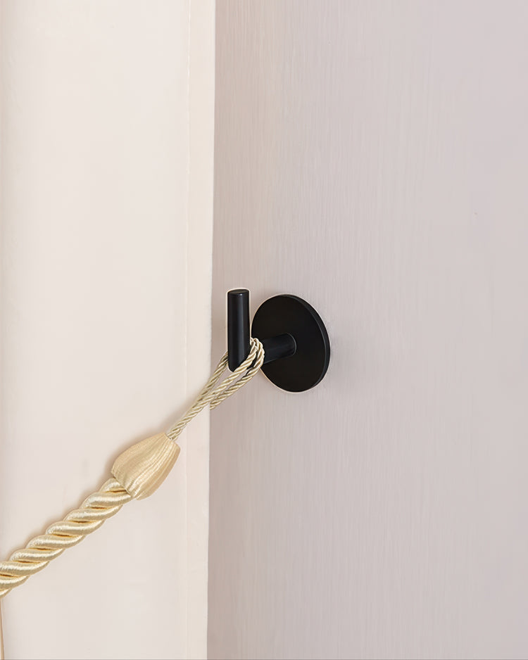 Sleek black modern curtain wall hook holding a golden braided drapery tie-back against a light wall.