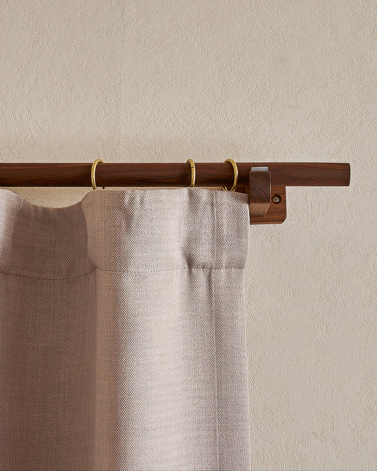 Minimal dark wood curtain rod with brass rings holding a tailored light beige linen drapery panel against a textured wall.