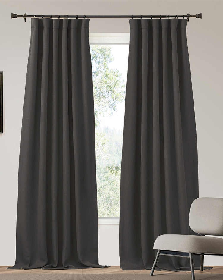 Zoe charcoal linen look heavyweight custom blackout drapery panels, partially open, hanging from a rod, with a glimpse of nature outside.