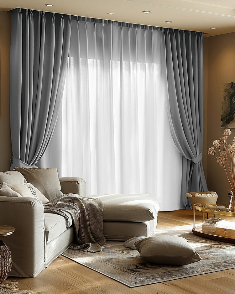 Sophisticated grey blackout velvet custom curtains with pleated headers, tied back, layered with sheer drapery in a bright living room.