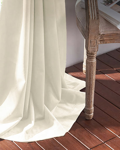 Cream 100% blackout velvet custom curtain, elegantly flowing onto dark wood floors beside a decorative carved chair leg.