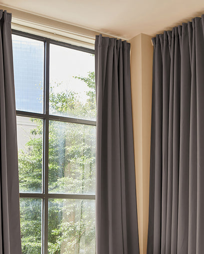 Hussein premium custom dark grey blackout drapery panels with elegant pleats, framing a sunlit window.