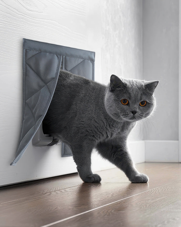 Grey cat with orange eyes exiting a grey quilted custom thermal pet door cover installed on a white door.