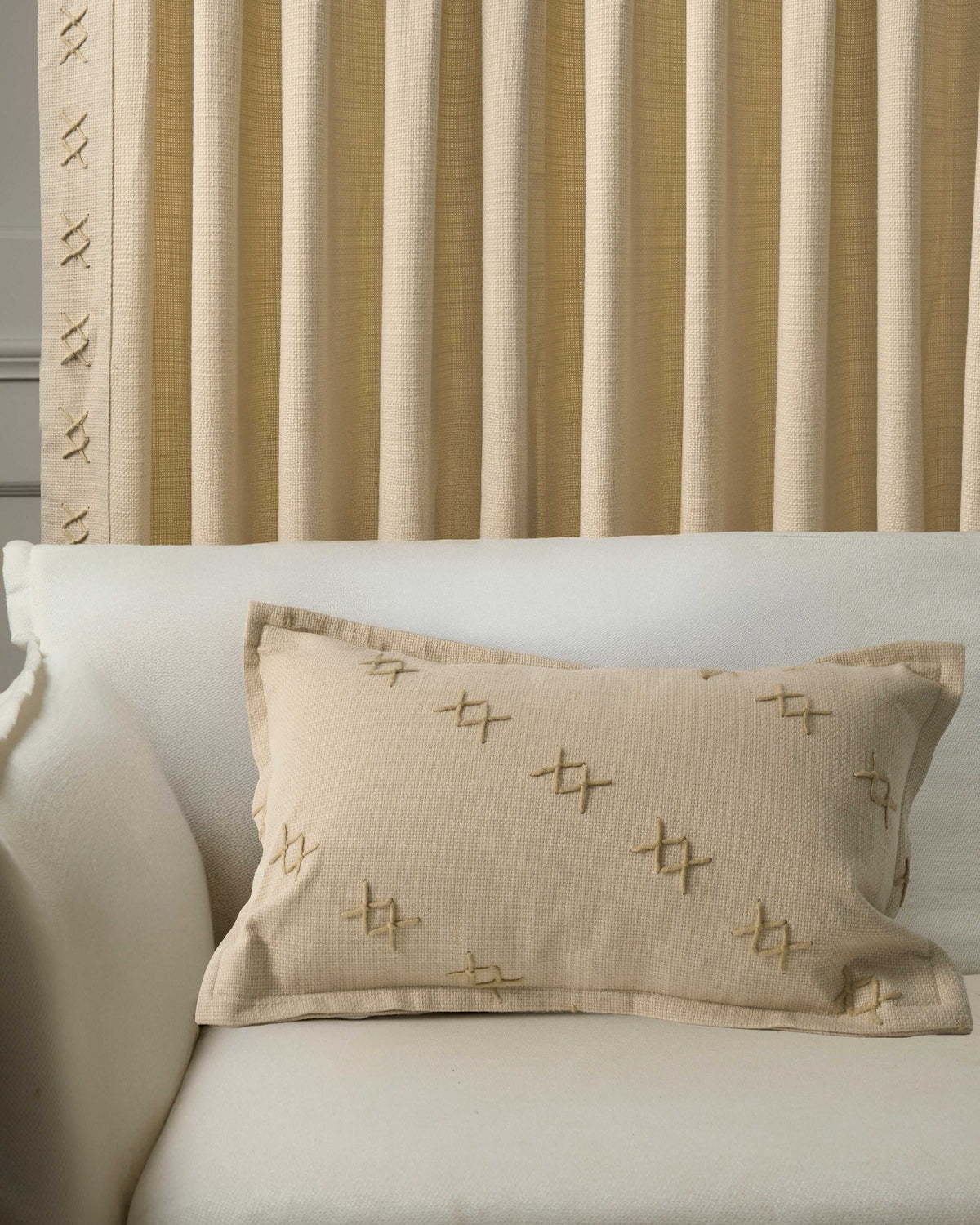 Arden textured lumbar pillowcase featuring decorative "x" yarn stitching on a white sofa, beside matching pleated window drapery.