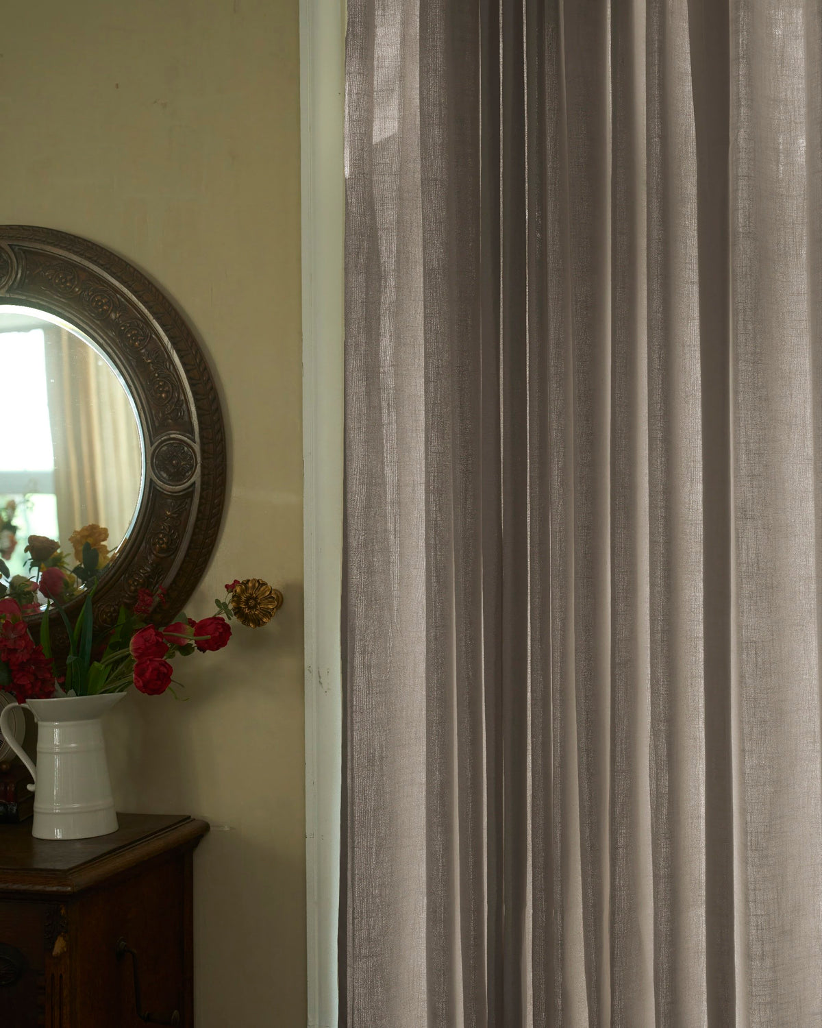Cassie linen blend custom sheer curtains, taupe textured fabric at a sunlit window. Ornate mirror & flowers on dark wood chest.