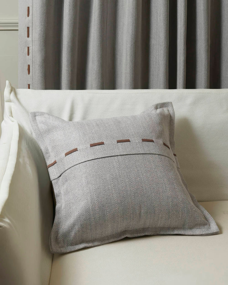 Cleo textured grey herringbone throw pillowcase with brown faux leather lacing, styled on a sofa, with grey drapery.