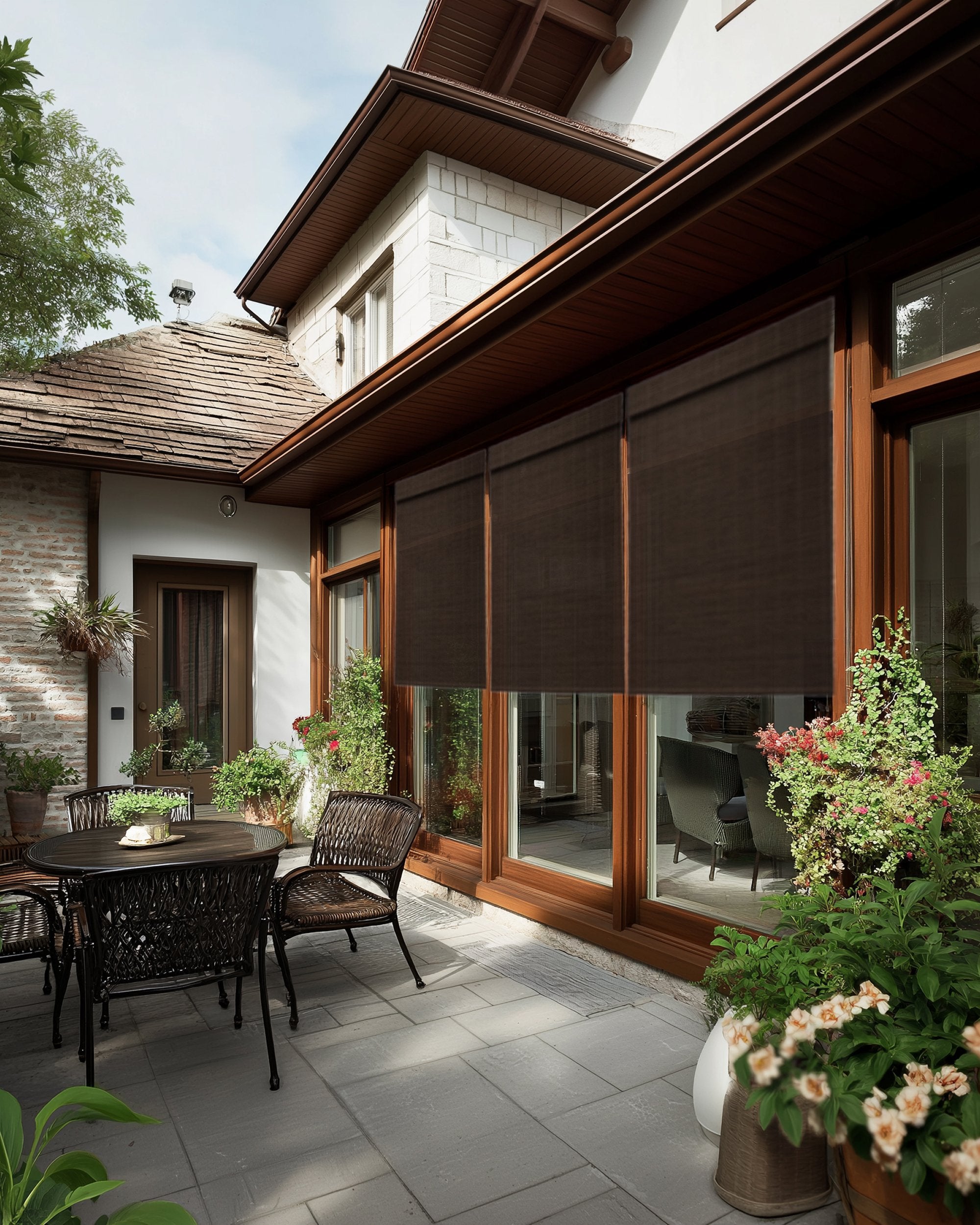 Hana Bark bamboo outdoor woven shades on large windows provide sun protection for a serene patio with outdoor furniture & lush plants.
