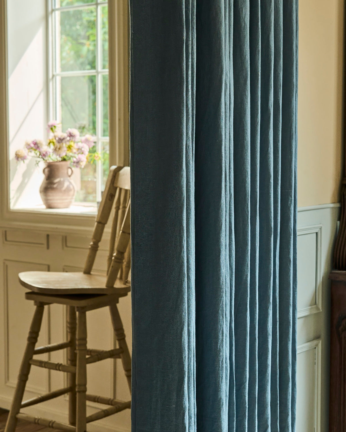 Muted blue Helena custom linen drapery, finely pleated, with sunlight softly filtering through a window and a rustic wooden chair.