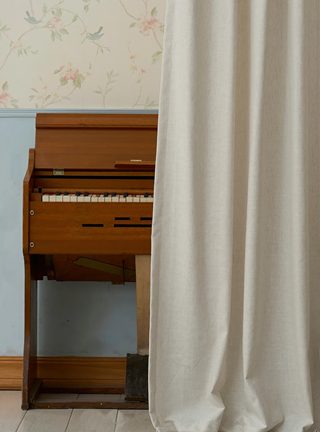 Wooden piano against a floral wallpapered wall with a white curtain.