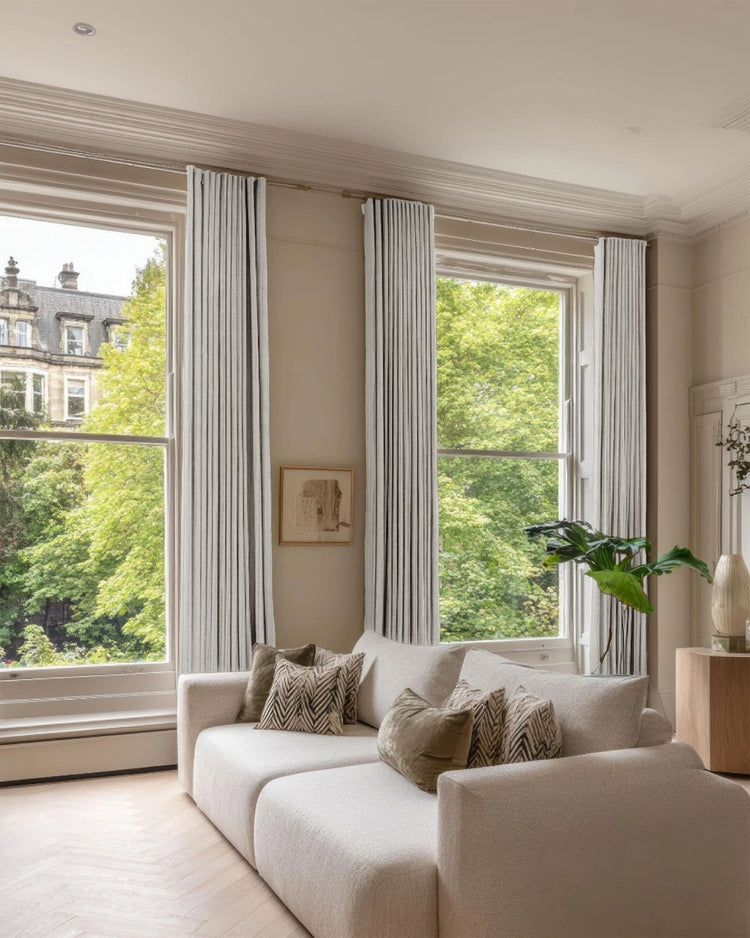 Sahara 100% blackout linen blend patterned custom drapery framing tall windows in a modern living room with a sofa & green outdoor view.