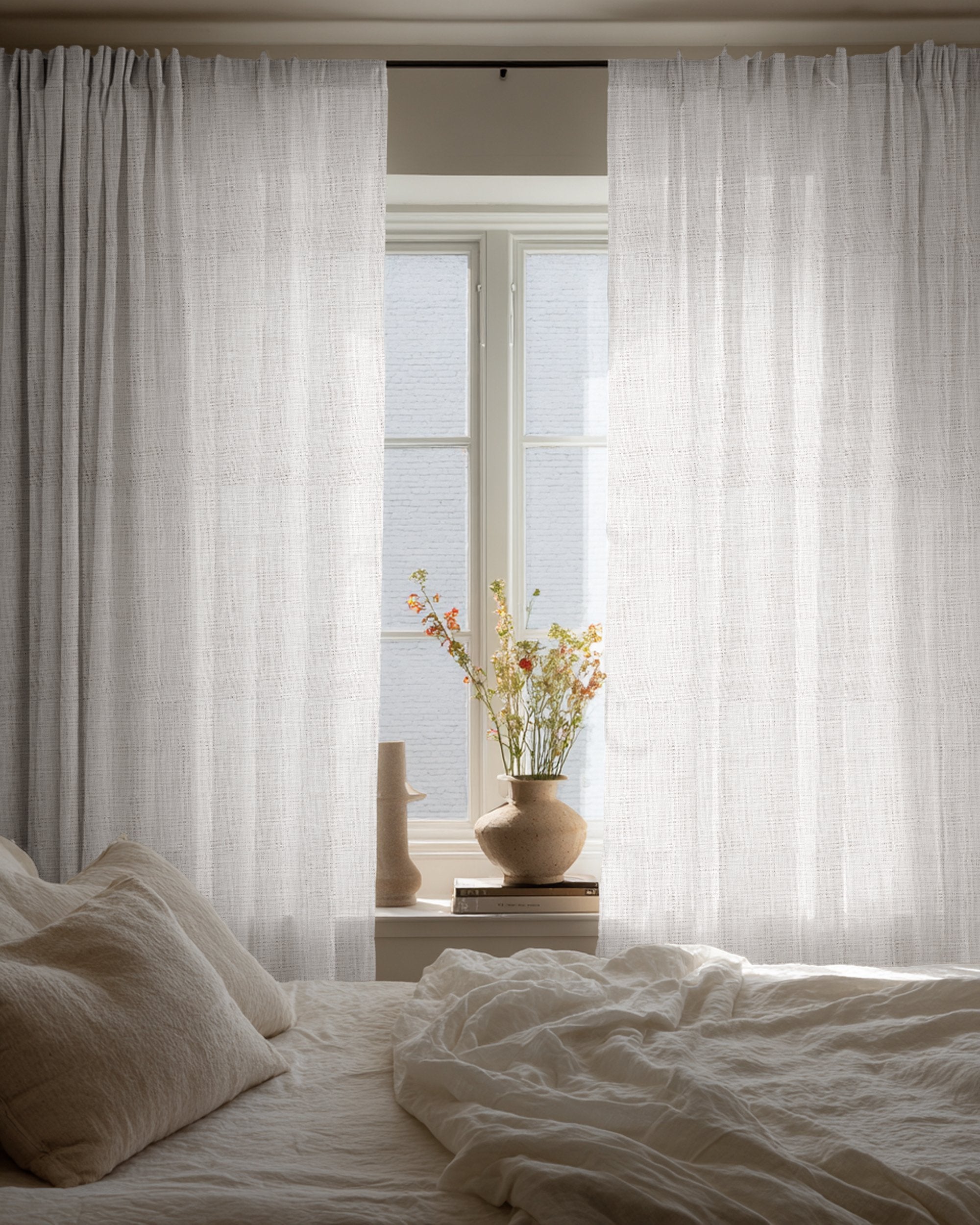 Custom Sunveil pure linen sheer curtains hanging by a bedroom window, diffusing natural light over a cozy bed and decorative vase.