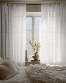 Custom Sunveil pure linen sheer curtains hanging by a bedroom window, diffusing natural light over a cozy bed and decorative vase.