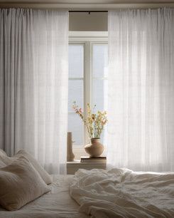 Custom Sunveil pure linen sheer curtains hanging by a bedroom window, diffusing natural light over a cozy bed and decorative vase.