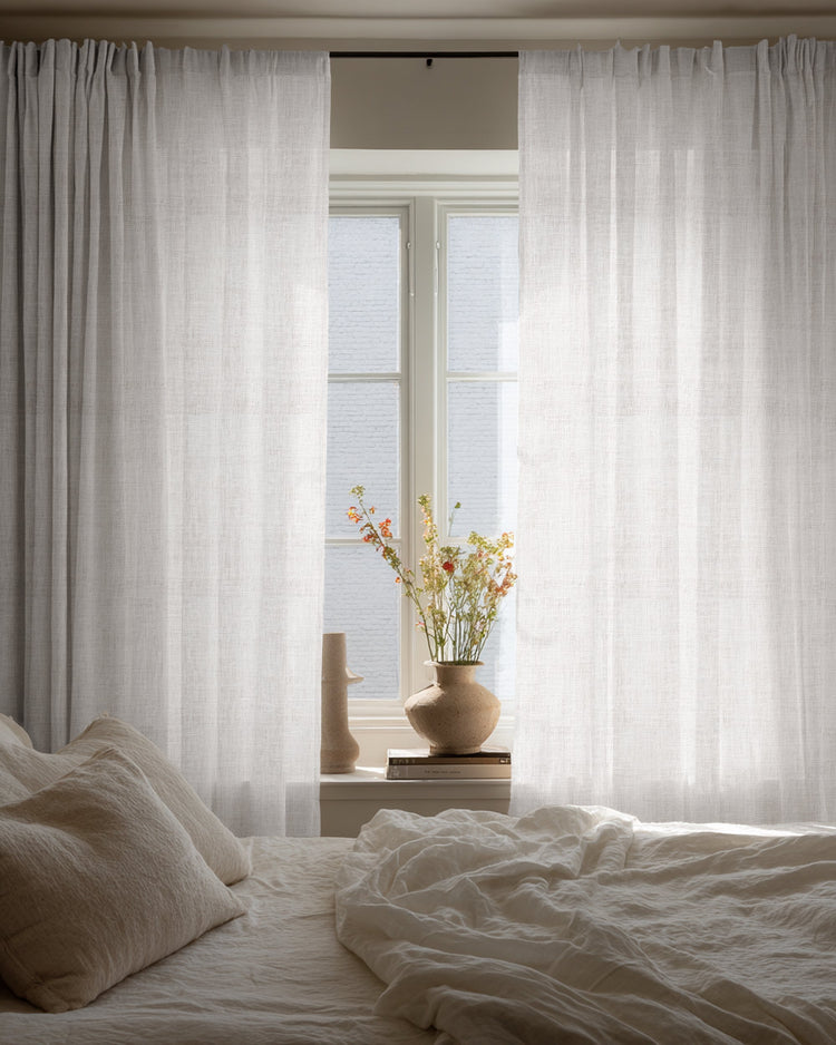 Custom Sunveil pure linen sheer curtains hanging by a bedroom window, diffusing natural light over a cozy bed and decorative vase.