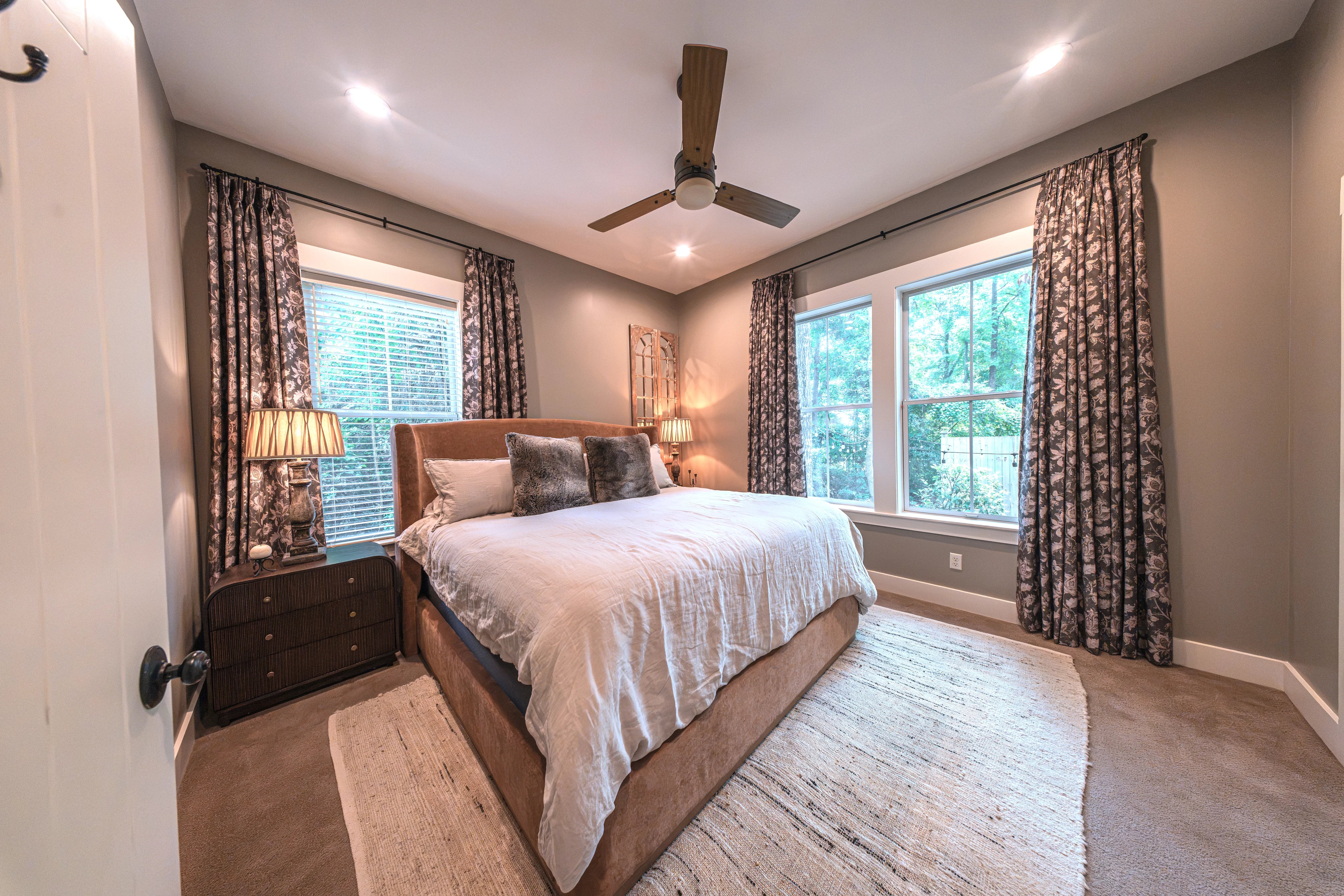 A styled bedroom interior featuring a bed with white bedding, a brown upholstered bed frame, patterned curtains, nightstands with lamps, and a ceiling fan on a neutral gray wall.