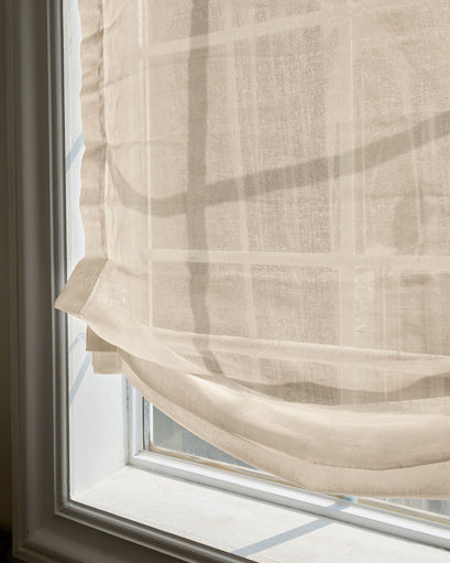 Light beige yarn sheer relaxed custom Roman shade on a white window, with sunlight filtering through its delicate fabric texture and soft folds.