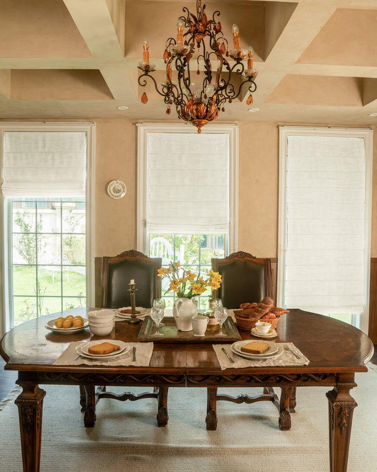 Dining room with three windows featuring light cream custom Zoe Plain Fold linen-look Roman shades, providing elegant light control.