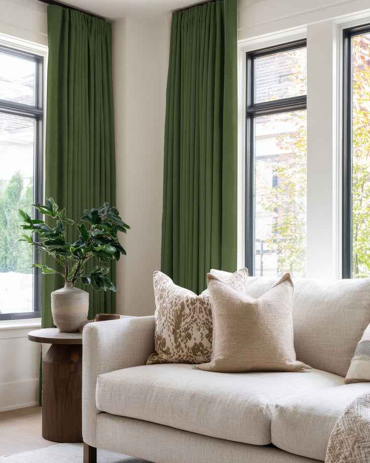 Dark green linen look blackout custom curtains adorning large windows in a bright living room with a neutral sofa and plant.