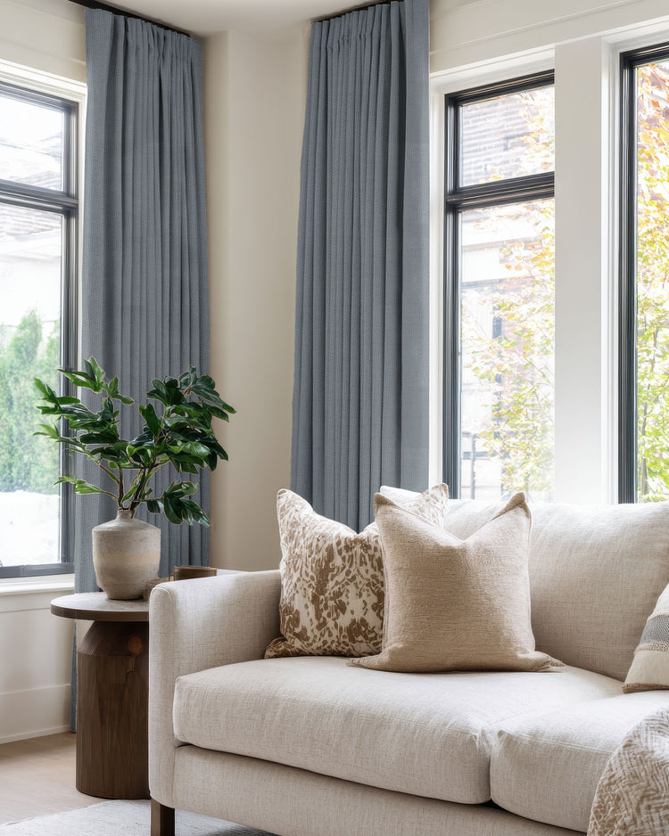Zoe 100% blackout linen look custom grey-blue pleated drapery on tall living room windows, paired with a light sofa.