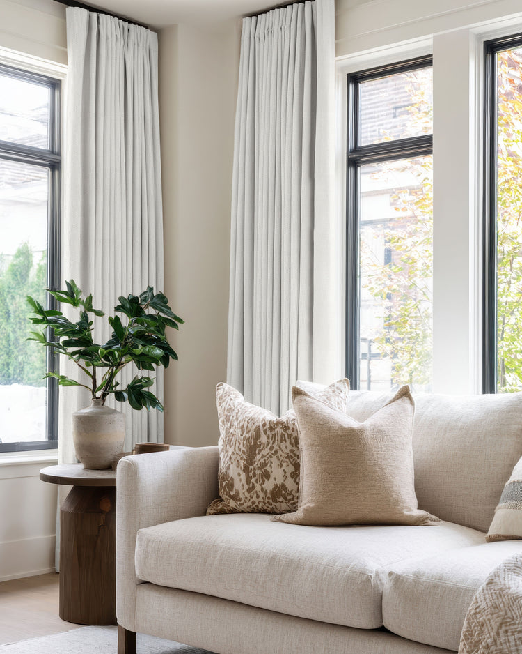 Zoe 100% Blackout Linen Look custom curtains in light gray with pinch pleats, framing large windows in a bright living room with a cream sofa.